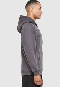 Sudadera gris con capucha, bolsillo tipo canguro, puños y dobladillo acanalados. Tejido suave con un ajuste holgado, mostrada de lado.