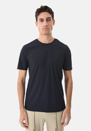 Uomo con capelli corti e scuri che indossa una maglietta nera a maniche corte semplice e pantaloni beige, in piedi davanti a uno sfondo bianco.