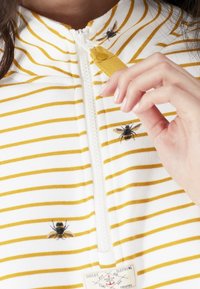 Prenda blanca con cremallera y rayas amarillas, que presenta abejas bordadas y una lengüeta amarilla de la cremallera. La tela parece suave y con textura.