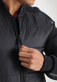 Veste noire matelassée avec un devant zippé, présentant des manches en tissu doux, un col côtelé et de subtils accents réfléchissants sur les épaules.