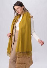 Sciarpa gialla senape con bordi frangiati, realizzata in un tessuto morbido; indossata con una gonna fitted beige e un gilet imbottito color tan.