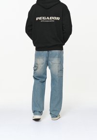 Svart hoodie med texten "PEGAJOR" och trycket "CERTIFIED AUTHENTIC STREETWEAR". Blå, löst sittande jeans med fickor och blekt utseende.