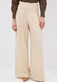 Pantaloni beige a gamba larga con vita alta, pieghe frontali e una texture liscia, abbinati a tacchi di colore chiaro.