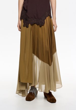 Falda larga y con capas en tonos terrenales de marrón y beige; cuenta con un tejido transparente, un dobladillo desigual y una silueta fluida. Combinada con zapatos marrones.