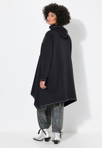 Zwarte capuchon poncho met een losse, asymmetrische vorm. Kenmerkt zich door contrasterende stiksels en is gecombineerd met glanzende, paillettenbroek en zilveren laarzen.