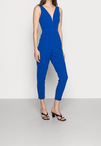 Mono azul sin mangas con escote en V profundo, piernas estrechas y bolsillos laterales, combinado con sandalias negras de tacón ancho. Textura suave.