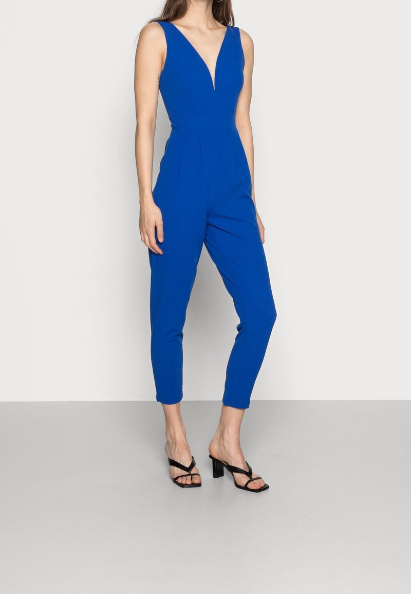 Mono azul sin mangas con escote en V profundo, piernas estrechas y bolsillos laterales, combinado con sandalias negras de tacón ancho. Textura suave.