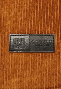 Brunt kordflorsmaterial har vertikal ribb; en svart läderetikett visar DC:s logga och texten "KVALITETSPRODUKTER SEDAN 1994."