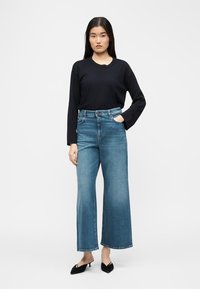 Γυναίκα με μαύρο πουκάμισο μακριά μανίκια, ψηλόμεσα φαρδιά μπλε τζιν παντελόνια και μαύρα μυτερά τακούνια, στέκεται μπροστά σε λευκό φόντο.