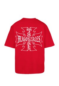 Κόκκινο βαμβακερό T-shirt με άνετη εφαρμογή, με μεγάλο λευκό γραφικό σχέδιο ενός σταυρού και το κείμενο "MIGONZALES" στην πλάτη.