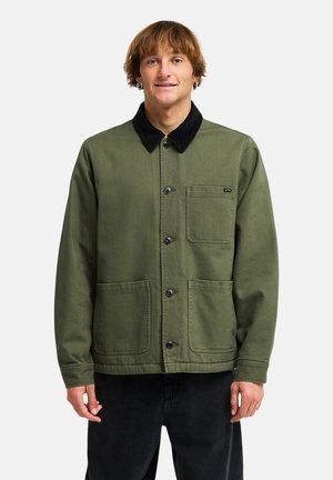 Jeune homme aux cheveux courts châtain clair portant une veste verte boutonnée avec un col noir et trois poches avant, debout sur fond blanc.
