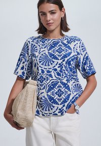 Sinisellä ja valkoisella kuviolla koristeltu lyhythihainen paita rentolla istuvuudella, valmistettu kevyestä kankaasta, yhdistetty teksturoituun beigen väriseen laukkuun ja valkoisiin housuihin.