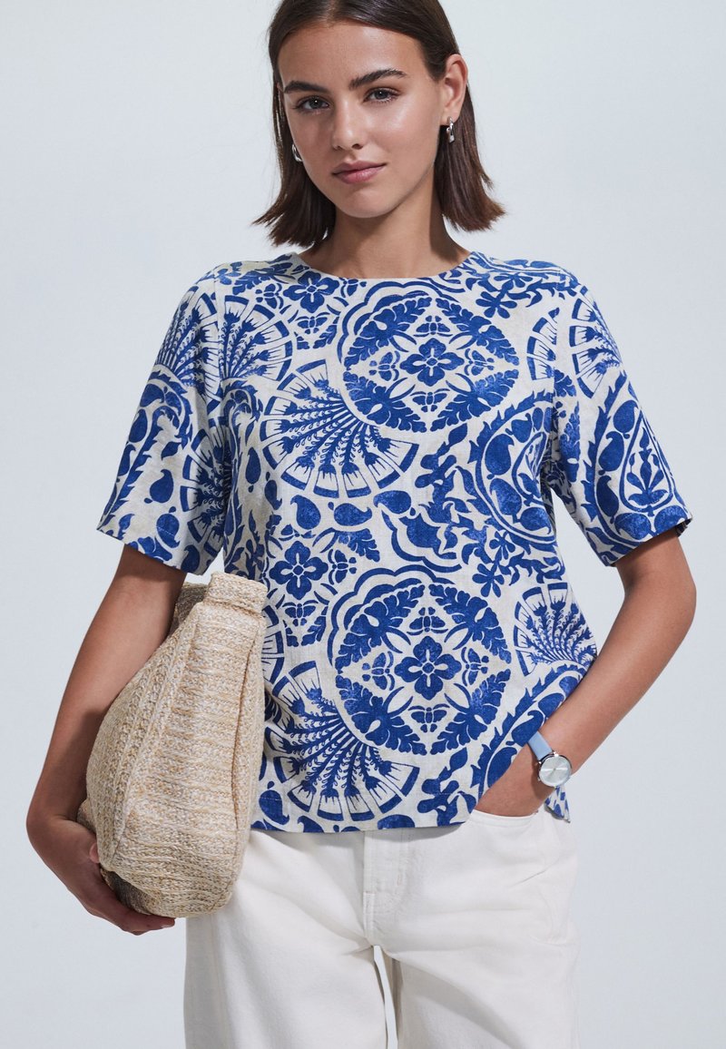 Sinisellä ja valkoisella kuviolla koristeltu lyhythihainen paita rentolla istuvuudella, valmistettu kevyestä kankaasta, yhdistetty teksturoituun beigen väriseen laukkuun ja valkoisiin housuihin.