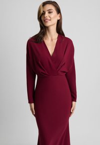 Burgundowa sukienka z długim rękawem i dekoltem w kształcie litery V, z drapowaniami w okolicy biustu oraz dopasowanym, prostym krojem. Gładka tekstura materiału.