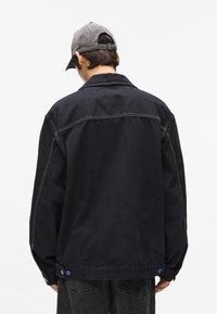 Mees, kes on seljaga kaamera poole, kannab musta jakk, millel on valged õmblused, musti teksaseid ja halli pesapallikiivrit, seistes ühetaolise valge tausta ees.