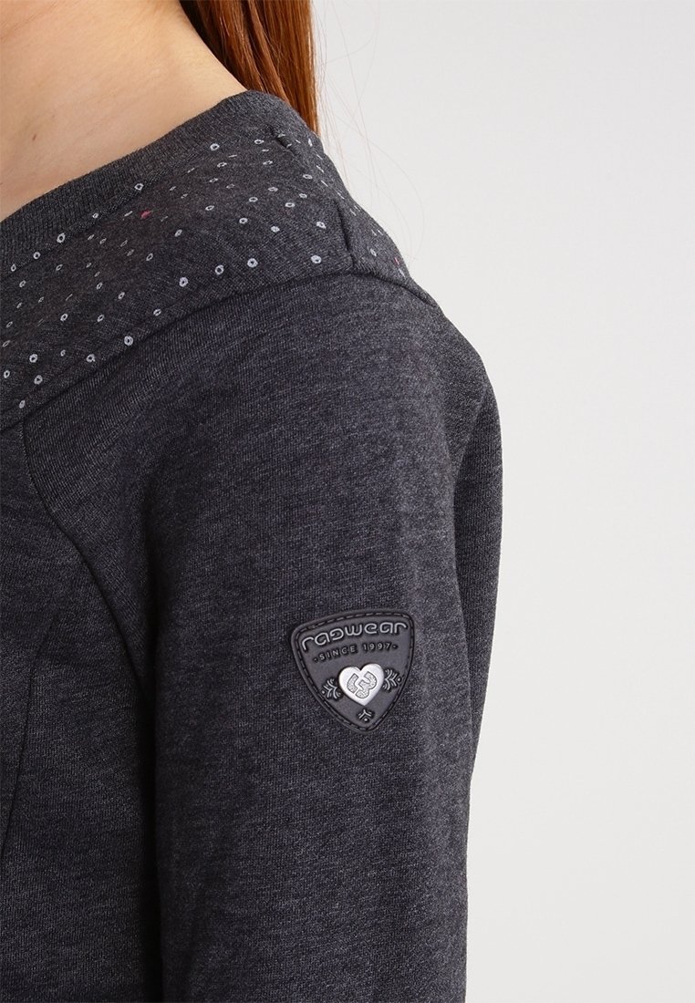 Gros plan sur une manche de sweatshirt gris foncé avec un col à motifs et un patch triangulaire « ragwear since 1997 » sur le haut du bras.