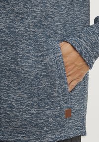 Mėlynas tekstūruotas audinio megztinis su priekiu kišene ir maža ruda odinė etikete apačioje. Dizainas pasižymi melange raštu.