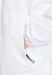 Biela bunda so zipsovou kapsou a čiernym ťahákom a elastickými manžetami. Tkanina je hladká s jemným leskom.