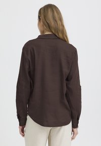Femme aux longs cheveux blonds portant une chemise à manches longues marron foncé et un pantalon beige clair, vue de dos sur un fond uni.