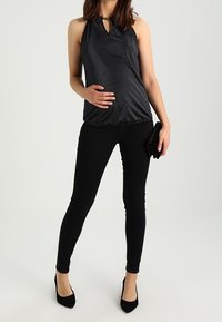 Svart halterneck-top med glänsande yta, nyckelhålsringning, matchat med svarta tajta byxor och svarta högklackade skor, hållande en liten kuvertväska.