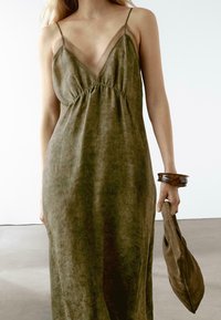 Olive green, sleeveless dress with a V-neck, gathered waist, and soft texture. Paired with a brown bangle and holding a matching bag.