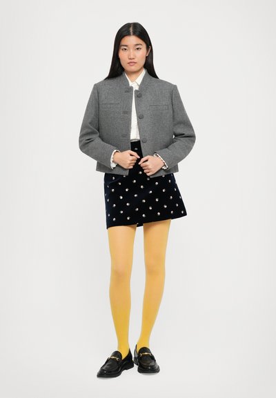 Žena stojící v šedém sako na knoflíky, bílém blouse s krajkovými manžetami, námořnické květinové sukni, žlutých punčochách a černých mokasínech se zlatým řetězem.