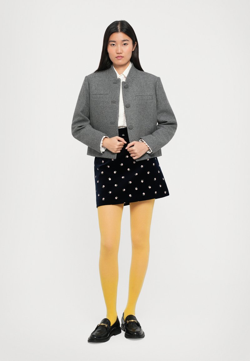 Femme debout portant une veste grise à boutons, un chemisier blanc avec des manchettes à volants, une jupe florale bleu marine, des collants jaunes et des mocassins noirs avec une chaîne en or.