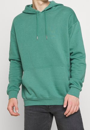 Homme portant un sweat à capuche vert avec poche avant et un jean noir, posant devant un fond clair uni.