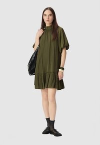 Vestido verde oliva con textura plisada, mangas cortas abullonadas y dobladillo con volantes. Combinado con zapatos negros y bolso negro.