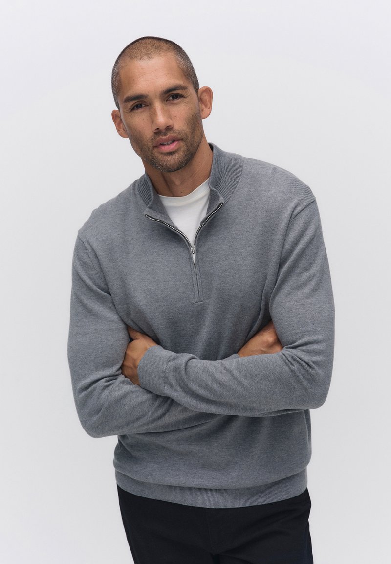 Grå zip-up sweatshirt med hög krage, ribbade ärmslut och nederkant, bärs över en vit skjorta. Modellen poserar med armarna i kors.