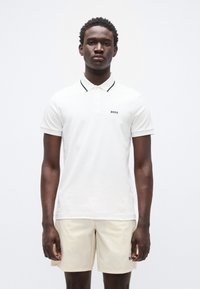 Λευκό πόλο μπλουζάκι με μαύρη επένδυση και λογότυπο στο αριστερό στήθος, συνδυασμένο με μπεζ σορτς. Υλικό βαμβακερό, κοντά μανίκια, κλασική εφαρμογή.