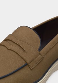 Mocassin en daim marron avec garniture noire et semelle blanche, présentant un détail de bride sur le dessus.