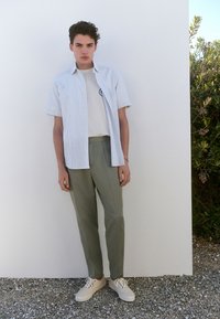 Chemise à manches courtes rayée bleu clair, t-shirt blanc en dessous, pantalon vert clair sur mesure, et chaussures beiges à lacets, debout sur du gravier.
