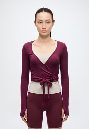 Une femme porte un haut cache-cœur bordeaux noué à la taille par-dessus une chemise crème, avec un short ajusté assorti, sur un fond uni.