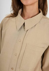 Camicia button-up beige realizzata in tessuto testurizzato. Caratterizzata da un colletto aperto, taschino frontale e bottoni beige. Design semplice e pulito.