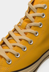 Κίτρινο καμβά ψηλό παπούτσι με μπεζ κορδόνια και μεταλλικά κορδόνια, με λαστιχένια σόλα και εμφανείς ραφές κατά μήκος των άκρων.