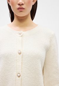 Crème kleurige, fluffy gebreide cardigan met een ronde halslijn, versierd met drie parelknopen met een goudkleurige rand langs de voorkant.