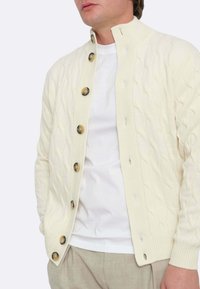 Cardigan lavorato a maglia color crema con motivo a treccia, scollo rotondo e grandi bottoni marroni, indossato sopra una camicia bianca. Tessuto dalla texture morbida.