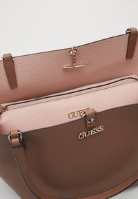 Sac à main en cuir texturé marron avec quincaillerie dorée, présentant un intérieur rose et un compartiment zippé. Logo de la marque visible.