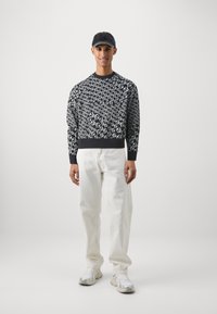 Svetr v námořnické modři s bílým abstraktním vzorem, doplněný volnými krémovými kalhotami a bílými sportovními teniskami. Stylizováno s tmavou čepicí.