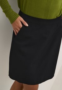 Black skirt with a smooth texture, featuring a flat waistband and side pocket. Pairing with a ribbed green long-sleeve top visible in the image.