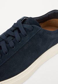Navy mokkasiner i suede med flade snørebånd, en rund tå og en kontrasterende lys cremet gummisål. Branding synlig på siden.