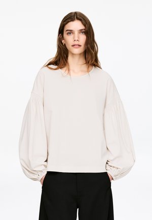 Lysebeige bluse med ballonsærmer, lavet af blødt stof. Har rund halsudskæring og elastiske manchetter. Kombinationen er med sorte bukser.