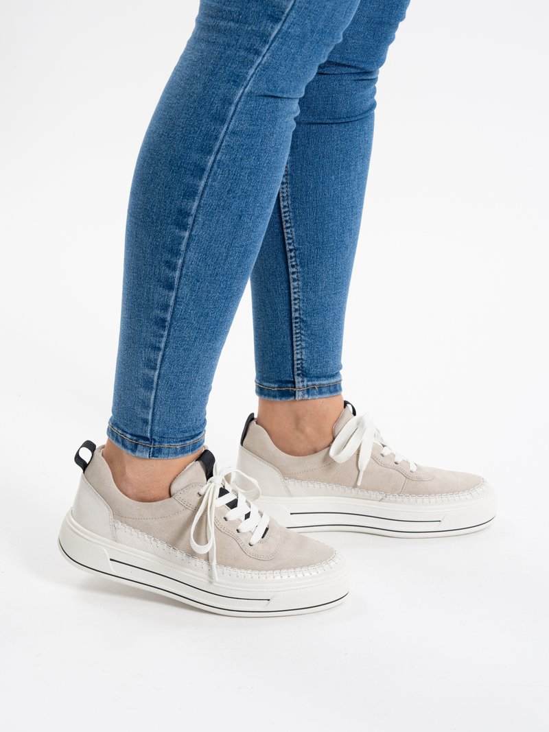 Legs wearing slim blue jeans and light beige lace-up sneakers with thick white soles and black pull tabs against white background.