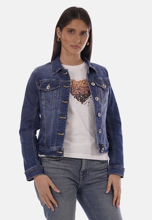 Donna che indossa una giacca di jeans blu sopra una t-shirt bianca con un disegno a cuore marrone, abbinata a jeans grigi, in piedi contro uno sfondo chiaro.