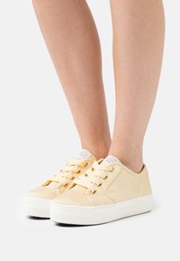 Gelbe Canvas-Sneaker mit einer weißen Gummisohle, runder Zehenpartie, Schnürdesign und Branding auf der Zunge. Glatte Textur.