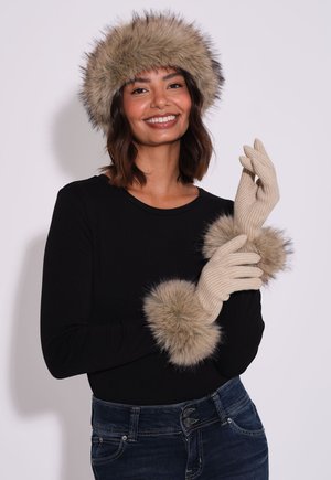 Frau lächelt, trägt ein schwarzes Top, blaue Jeans, beige Handschuhe und einen Hut mit Pelzbesatz und richtet einen Handschuh mit der anderen Hand.