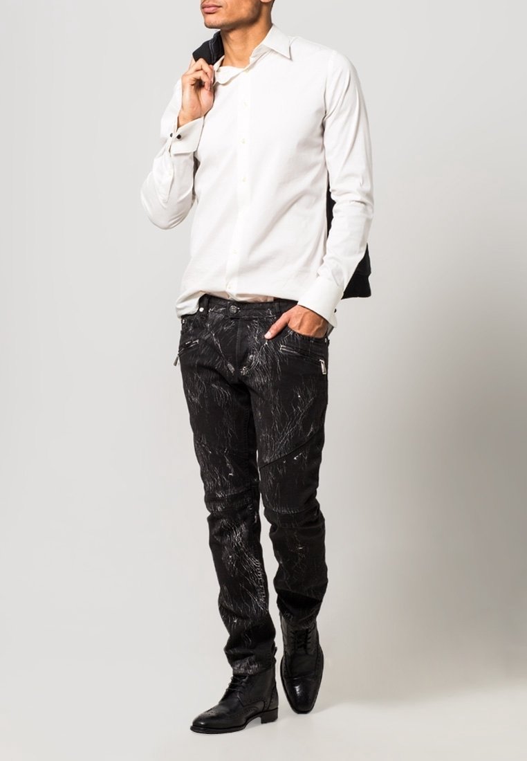 Camicia bianca con bottoni, jeans neri con motivi testurizzati, stivali di pelle nera e una giacca nera gettata sulla spalla.