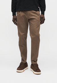 Pantalon en coton marron à coupe droite, avec des plis à l'avant et des poches latérales, associé à des baskets marron foncé.