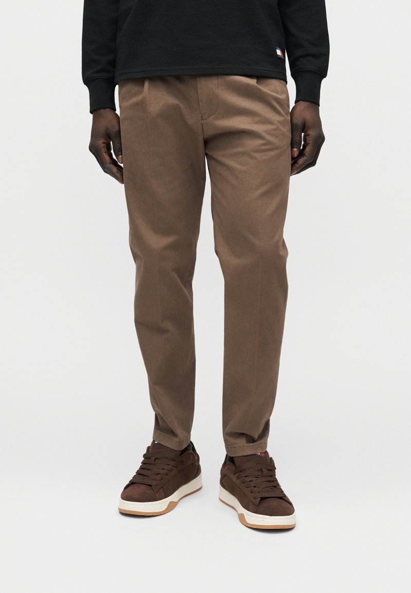 Pantalon en coton marron à coupe droite, avec des plis à l'avant et des poches latérales, associé à des baskets marron foncé.